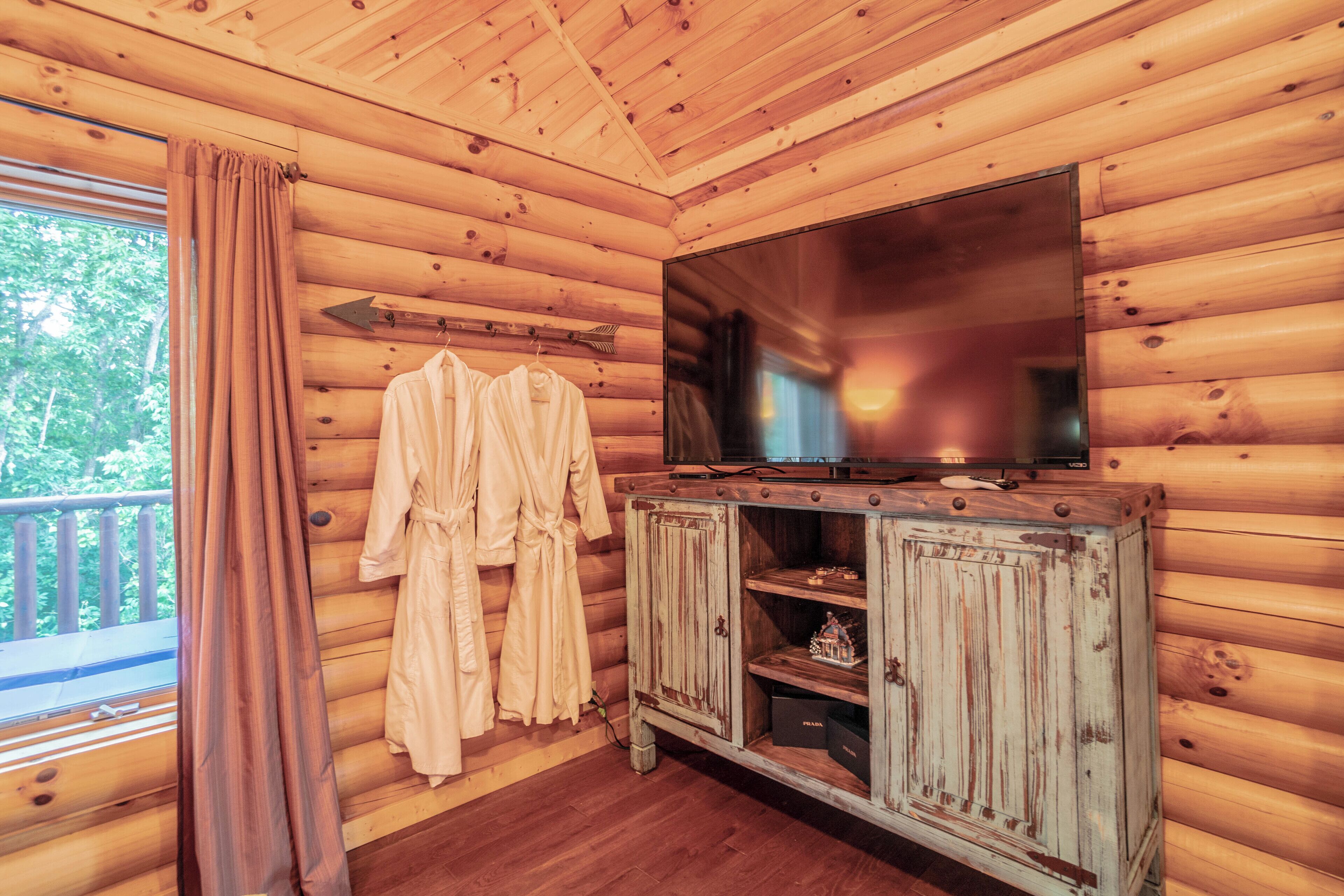 Ein Zimmer in einer Holzhütte mit einem großen Fernseher an der Wand, einem Schrank zur Aufbewahrung und einem Fenster mit Vorhängen.