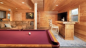 A wooden cabin interior with a pool table, a sofa, a dining area, and a TV.