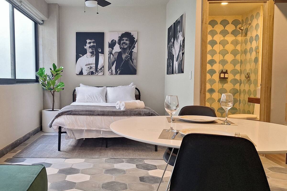 A hotel room with a bed, a dining table with chairs, a potted plant, and artwork on the walls.