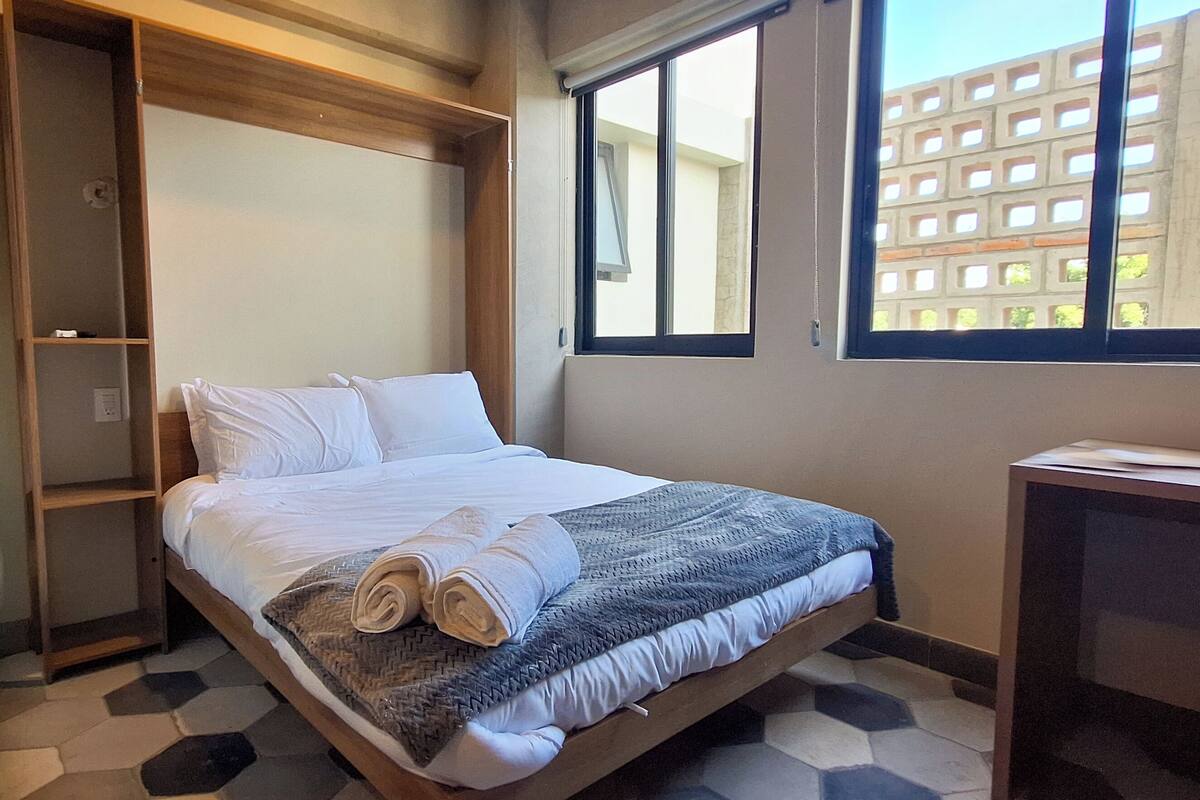 A hotel room with a bed, a desk, and a window with a view of buildings.