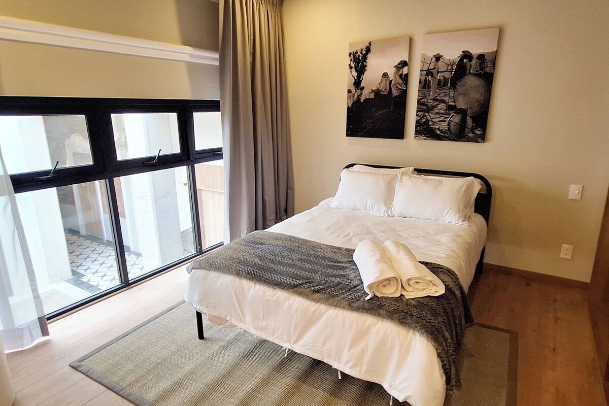 A bedroom with a bed, a large window, a plant, and artwork on the wall.