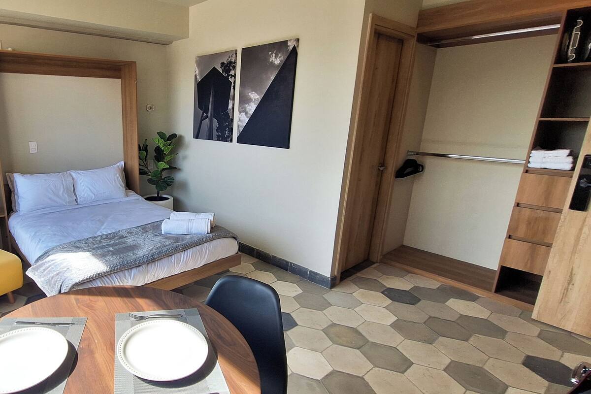 A compact room with a bed, a yellow armchair, a dining table with plates, and a potted plant.