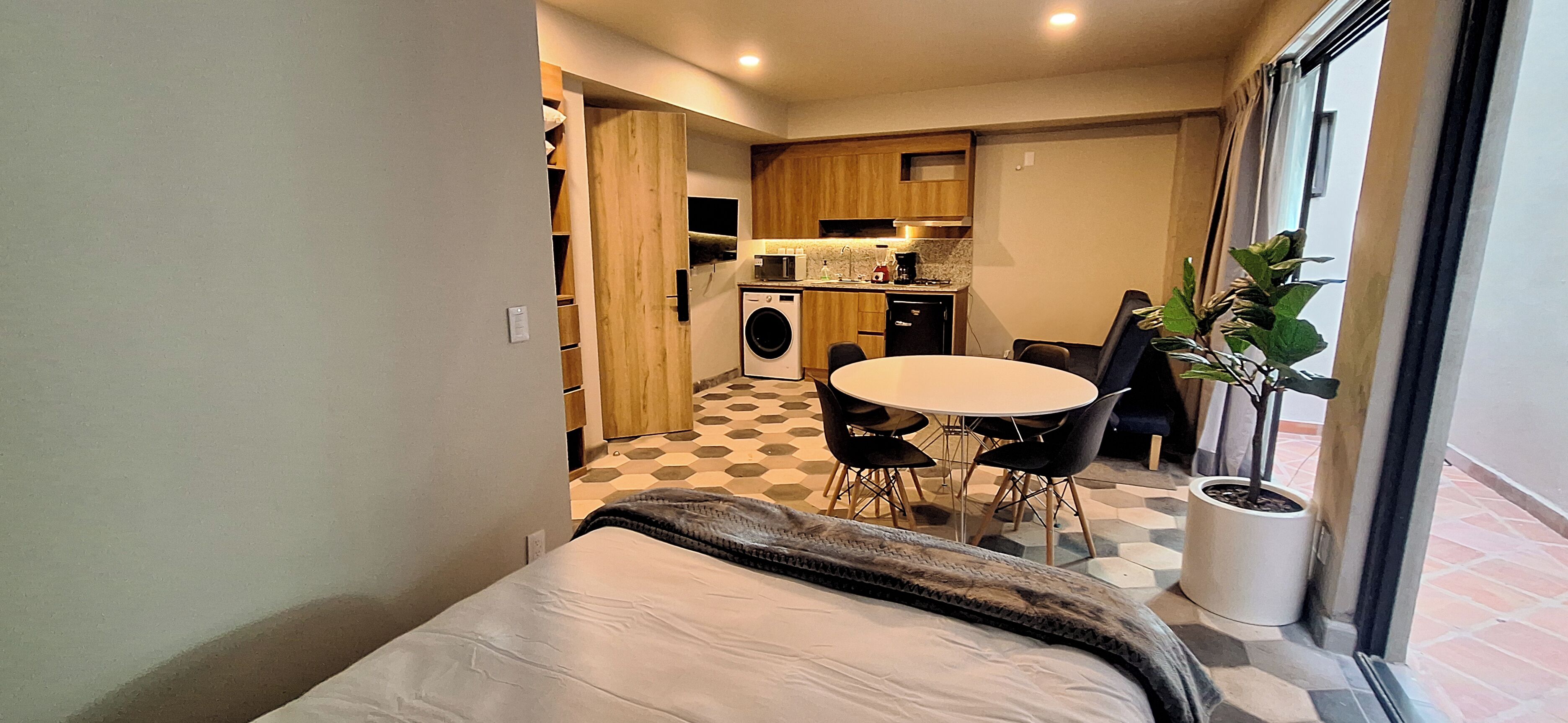 Een compacte hotelkamer met een bed, een klein eettafeltje met stoelen, een wasmachine en een potplant bij het raam.