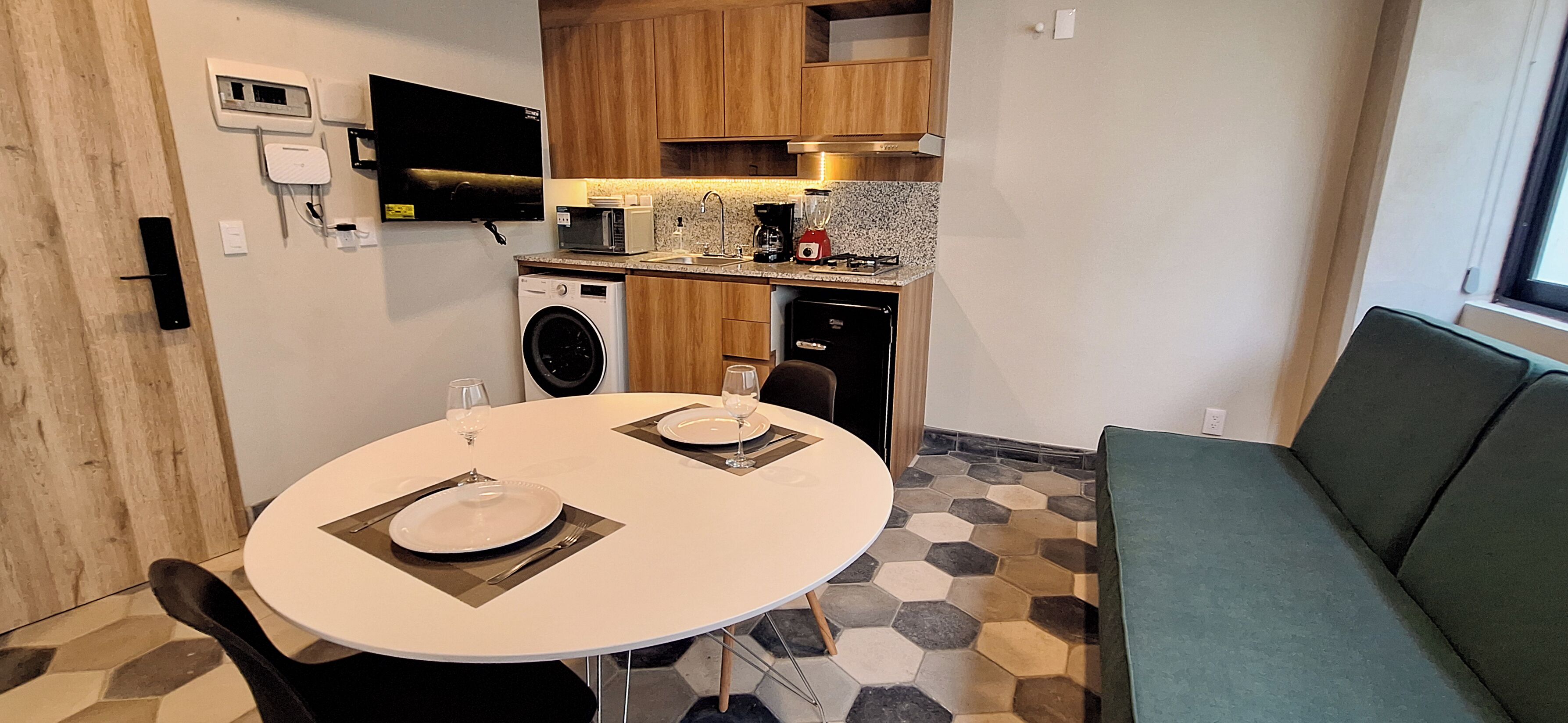 A compact kitchen with a washing machine, microwave, and coffee maker. There is a dining table with two plates and cutlery. A green sofa is visible in the foreground.