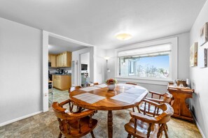 A dining area with a round wooden table, six chairs, and a vase with flowers. There is a window with a view of trees and a building outside. - Gog and Pop's Beach House - Ocean Views, WiFi, BBQ (Trinidad)