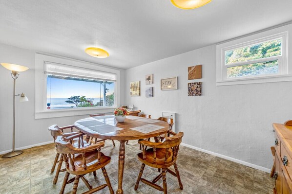 A dining area with a round wooden table and chairs, a view of the ocean through a window, and a ceiling light fixture.