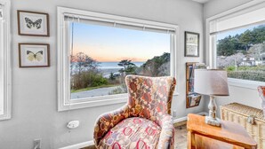 A room with a large window offering a view of the ocean and trees, a patterned armchair, a lamp, and framed artwork on the wall. - Gog and Pop's Beach House - Ocean Views, WiFi, BBQ (Trinidad)