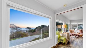 A room with a large window overlooking a road and trees, a dining area with chairs and a table, and a view of the ocean at sunset. - Gog and Pop's Beach House - Ocean Views, WiFi, BBQ (Trinidad)