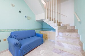 Een kamer met een blauwe bank, een trap met marmeren treden en een keukenhoek met een betegelde spatwand.