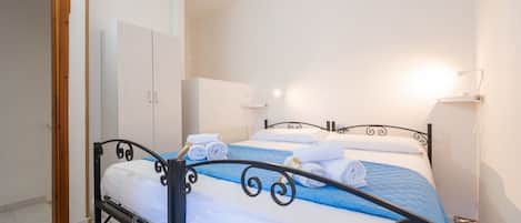 Una cama bien tendida con ropa de cama blanca y azul, un cabecero de metal negro y un armario blanco en una habitación sencilla con paredes blancas y una puerta de madera.