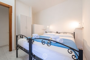 Ein ordentlich bezogenes Bett mit weißer und blauer Bettwäsche, ein schwarzer Metallrahmen und ein weißer Kleiderschrank in einem schlichten Raum mit weißen Wänden und einer Holztür.