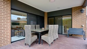 A covered patio with a table and chairs, a glass door leading to a garden, and a brick wall. - Point Cook Serenity Stay (Point Cook)