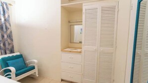A bedroom with a white bed, a blue chair, a white wardrobe with a mirror, and a bathroom with a shower curtain.