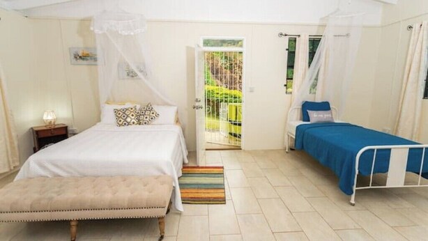 A bedroom with two beds, a bench, and a view of a garden through an open door.