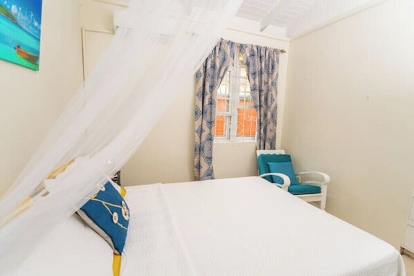 A bedroom with a bed, a chair, a window with curtains, and a painting on the wall. - Providence ,Charming 3-bedroom cottage with WiFi and AC in Mahaut. (Mahaut)