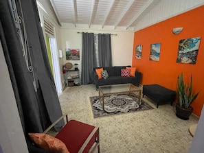 A living room with a sofa, coffee table, and a red chair. There is a large window with curtains, a potted plant, and a wall with paintings.