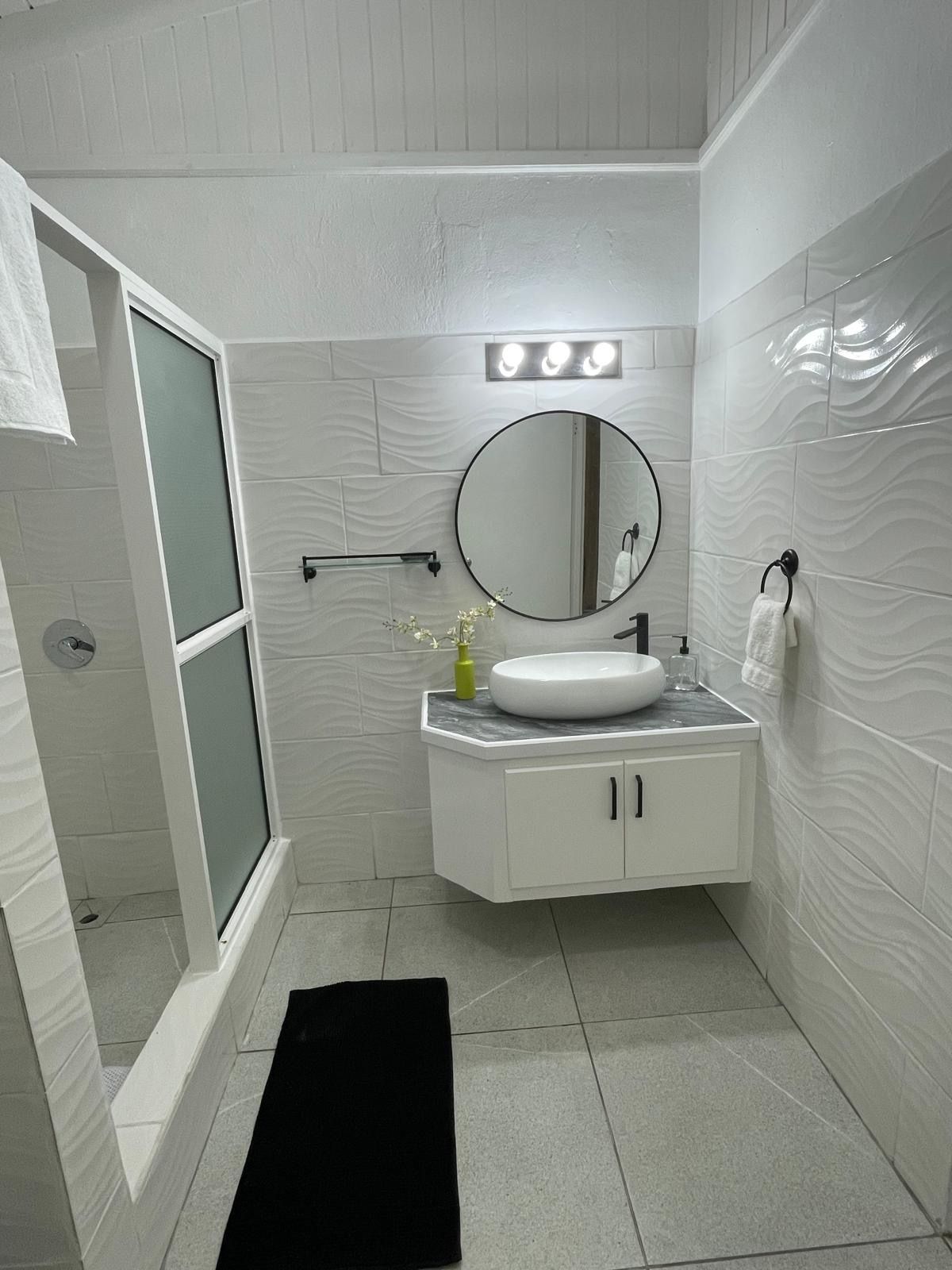 A bathroom with a white vanity, a round mirror, a white sink, and a glass shower enclosure.