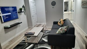 Una sala de estar moderna con un televisor de pantalla plana montado en la pared, un sofá gris con cojines estampados, una mesa de centro y una planta pequeña en un estante.