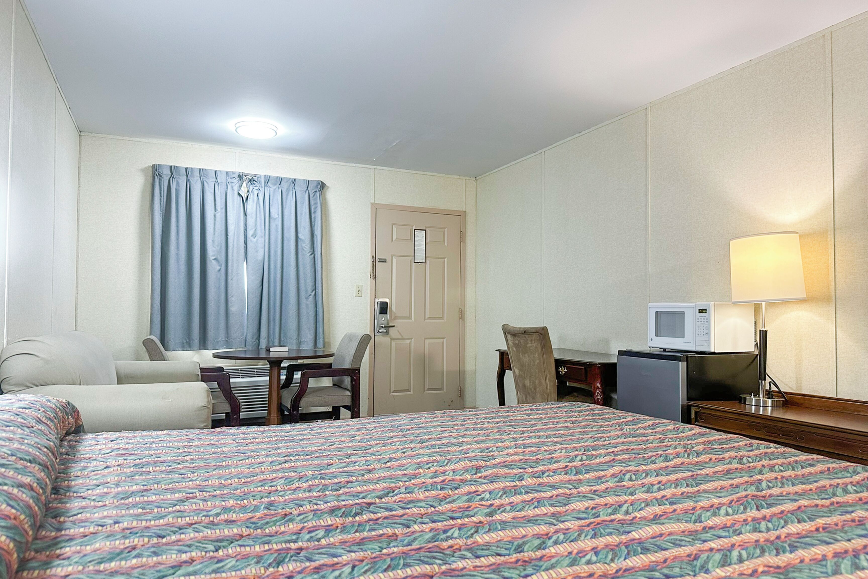 A hotel room with a large bed, a desk with a computer, a chair, a small table, a microwave, and a lamp.