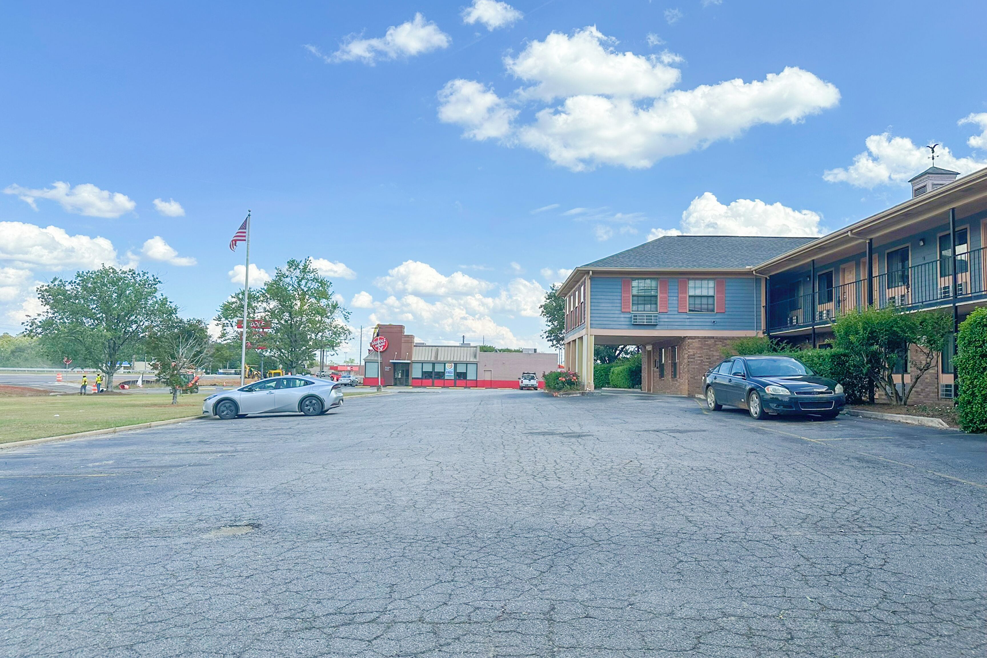 Egy parkoló, ahol két autó áll, egy vörös és kék színű homlokzatú épület, és egy amerikai zászló.