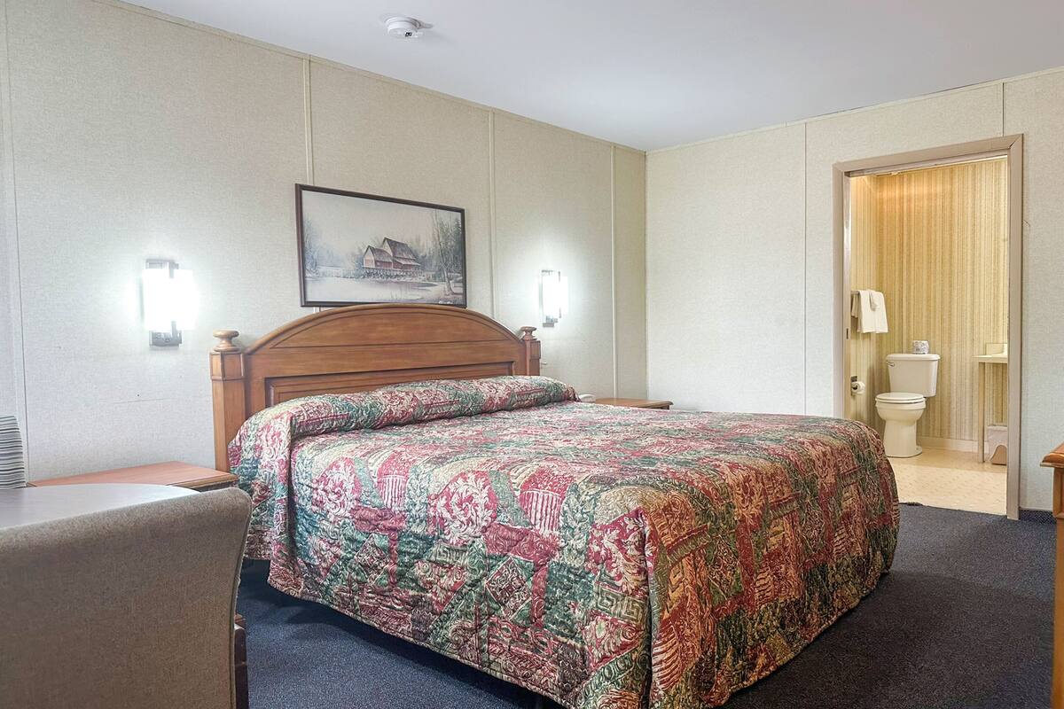 A hotel room with a bed, a wooden headboard, two wall-mounted lights, a framed picture, and a door leading to a bathroom.