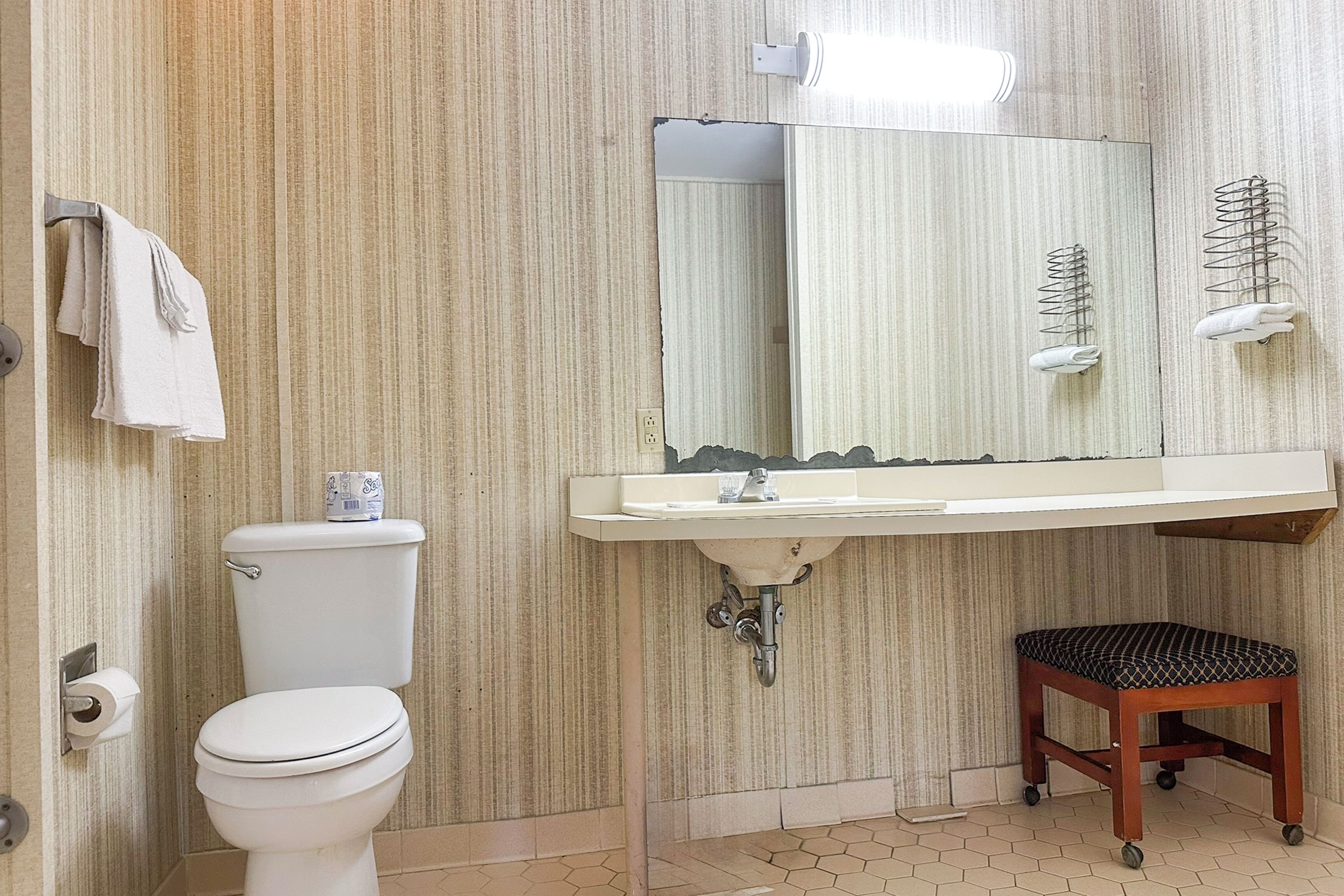 A bathroom with a toilet, sink, mirror, towel rack, and a small stool.