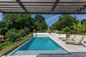 A rectangular swimming pool with a pergola overhead, surrounded by lush greenery and a few lounge chairs. - Villa Bichipau Park - Welkeys (Bidart)