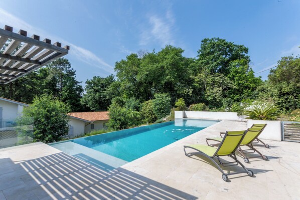 A swimming pool with clear blue water, surrounded by a patio area with two lounge chairs and a pergola. - Villa Bichipau Park - Welkeys (Bidart)