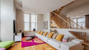Ein modernes Wohnzimmer mit einem weißen Sofa, bunten Kissen, einer Holztreppe und einem großen Fenster mit Jalousien.