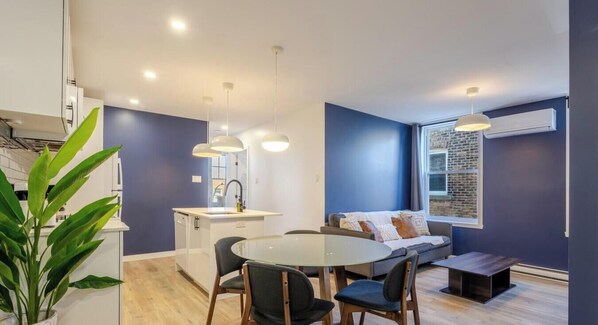 Una cocina moderna con armarios blancos, un comedor con mesa y sillas de vidrio, y una pared azul de acento.