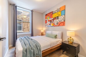 Une chambre à coucher avec un lit, une fenêtre avec des rideaux, un tableau décoratif au mur, une lampe de chevet et une table de chevet.
