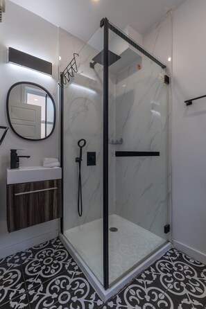 A modern bathroom with a glass-enclosed shower, a round mirror, a white sink, and patterned floor tiles. - Beautiful 3BR in Plateau Mont-Royal (Montréal)