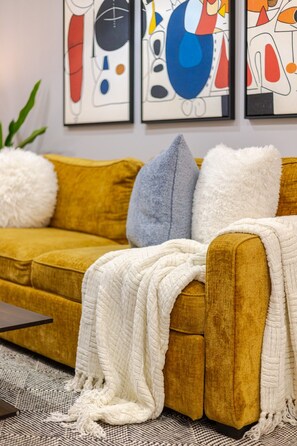 A mustard-colored sofa with a white throw blanket and two pillows, one white and one gray, in a room with abstract artwork on the wall. - Beautiful 3BR in Plateau Mont-Royal (Montréal)