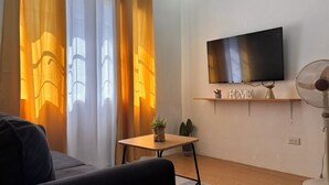 Ein Wohnzimmer mit einer Couch, einem an der Wand montierten Fernseher, einem kleinen Tisch mit einer Pflanze und einem Ventilator.