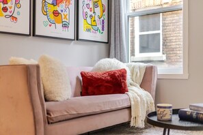 Un salon chaleureux avec un canapé, un coussin rouge et un plaid blanc. Des œuvres d’art encadrées ornent le mur, et une fenêtre offre une vue sur un bâtiment en briques.