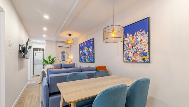 A modern dining area with a wooden table and blue chairs, a sofa, and two wall-mounted pendant lights. - Stunning 3BR in Plateau Mont-Royal (Montréal)