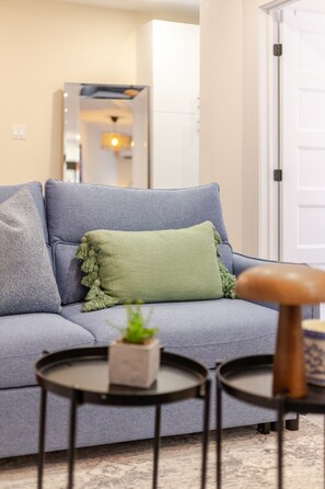 A living room with a grey sofa, a green throw pillow, a small table with a plant, and a wooden stool.
