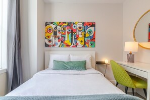 A hotel room with a bed, a desk, a chair, a lamp, and a colorful abstract painting on the wall.