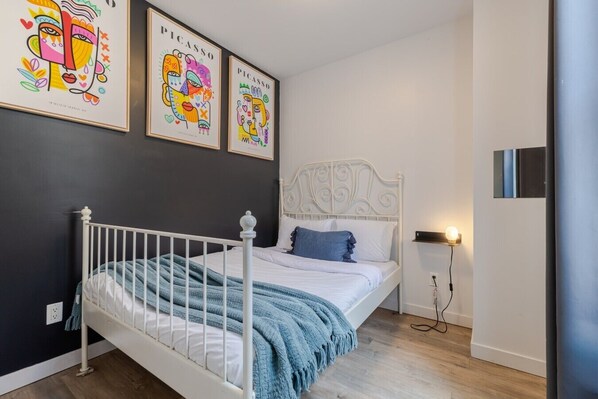 Un dormitorio con estructura de cama de metal blanco, manta azul y dos almohadas. Hay tres obras de arte enmarcadas en la pared sobre la cama.