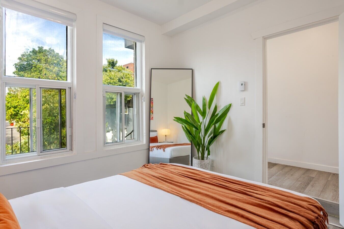 Une chambre à coucher comprenant un lit, une table de chevet, un miroir, une plante en pot et une fenêtre donnant sur des arbres et des immeubles.