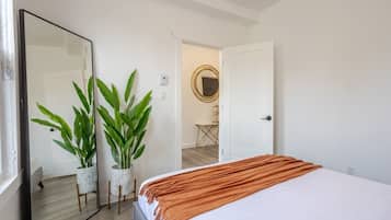 A bedroom with a bed, a large mirror, two potted plants, and a door leading to another room.