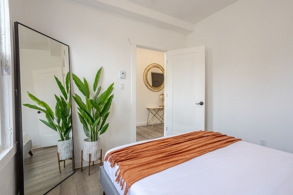 Une chambre à coucher avec un lit, un grand miroir, deux plantes en pot et une porte donnant sur une autre pièce.