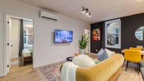 A modern living room with a flat-screen TV, a sofa, a dining area with yellow chairs, and a staircase leading to another level. - Vibrant 4BR/2BA Retreat in the Plateau (Montréal)
