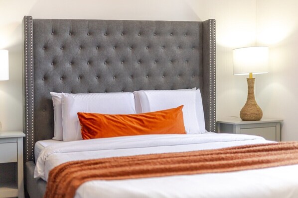 A hotel room with a tufted headboard, white bedding, an orange accent pillow, a striped throw blanket, and a bedside lamp.