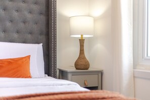 A bed with white and orange bedding, a grey upholstered headboard, a bedside table with a lamp, and a window with curtains.