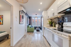 A modern kitchen with white cabinets, a stove, and a sink. There is a green sofa in the background and a window with a view of buildings. - The Perfect Downtown Hideaway (Montréal)