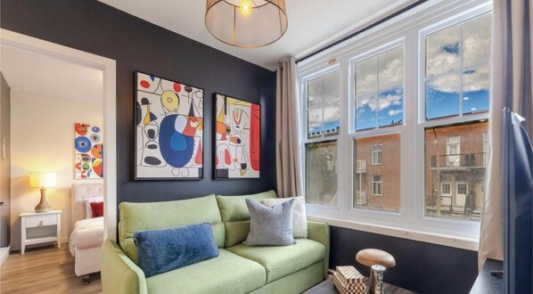 A living room with a green sofa, artwork on the wall, a lamp, and a view of buildings through large windows.