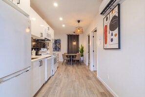 Un pasillo estrecho con una cocina a la izquierda, una zona de comedor con una mesa y sillas en el centro, y un cuadro abstracto enmarcado en la pared derecha.