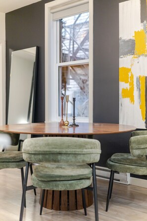 Een eetruimte met een ronde tafel, twee groene bekleden stoelen, een raam met uitzicht op een gebouw en een groot abstract schilderij aan de muur.