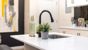 Una cocina con encimera blanca, grifo negro, una pequeña planta en maceta y una botella decorativa.